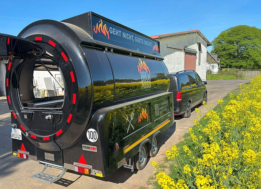 Schwarzer BBQ-Foodtruck mit Anhänger vor gelbem Rapsfeld.