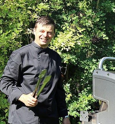 Grillmeister im schwarzen Outfit mit Grillzange am Gasgrill im sonnigen Garten