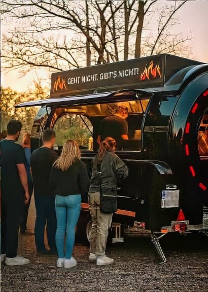Menschen genießen Bratwürste vor Foodtruck mit Schriftzug 'Geht nicht, gibts nicht' bei Grill-Event im Freien