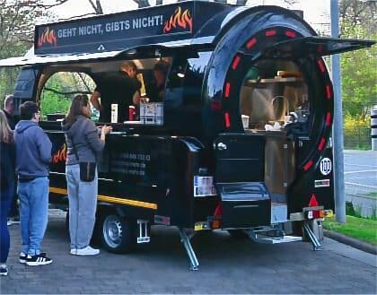 Schwarzer Foodtruck mit Flammenmotiv und Slogan „Geht nicht, gibt's nicht“ serviert Essen an wartende Kunden im Freien.