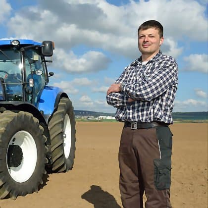 Landwirt steht auf Acker neben blauem Traktor unter blauem Himmel mit Wolken, bereit zur Feldarbeit
