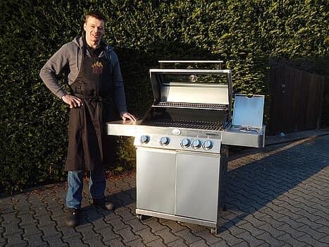 Mann mit Schürze neben Gasgrill auf gepflastertem Platz.